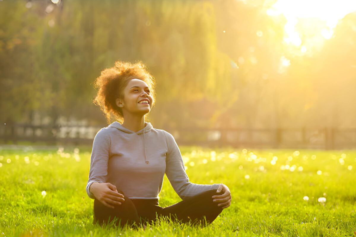 A woman practices creative self-care, smiling peacefully while basking in the sun in a grassy field.