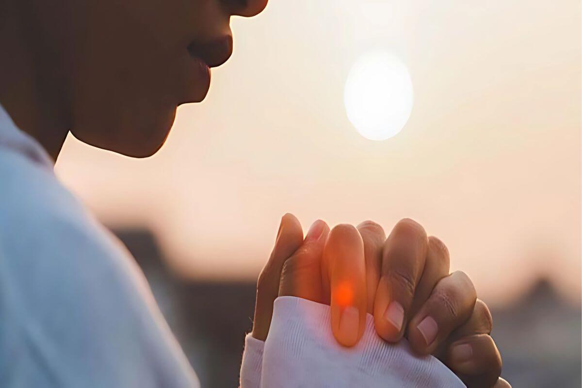 A person practices spiritual self-care, clasping their hands in prayer with the setting sun in the background.