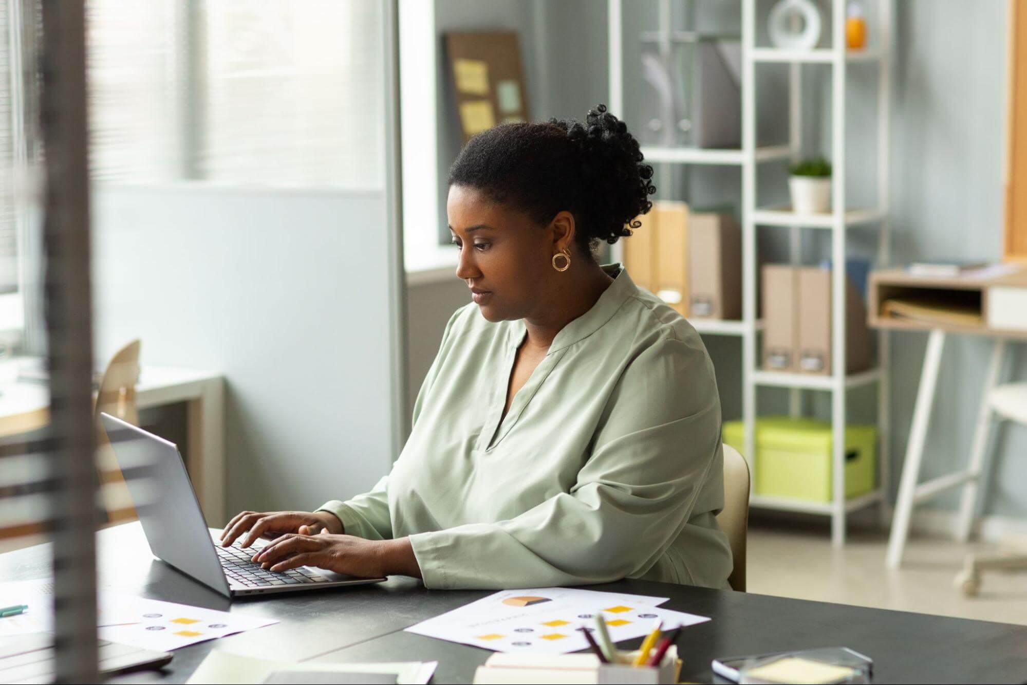 A professional woman with a bun works intently on her laptop at a desk, reflecting focus and strong work ethic.