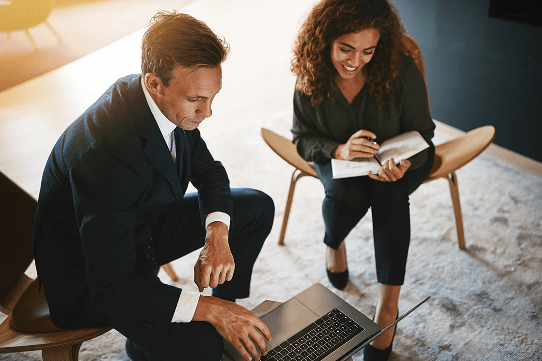 A business man mentoring a woman while they work together on a laptop and a notebook.