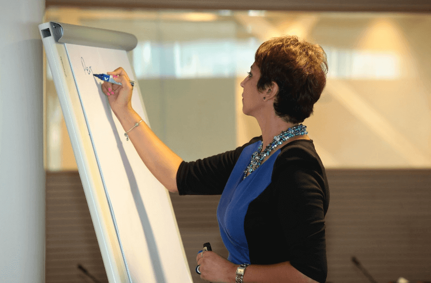 Mentor Leila Rezaiguia stands at a whiteboard, holding a blue marker and writing notes during a session.