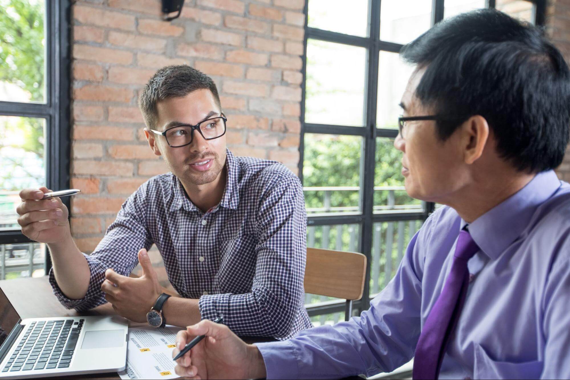  A mentor is speaking to a colleague at a table, gesturing while the other listens attentively with laptop and documents. 