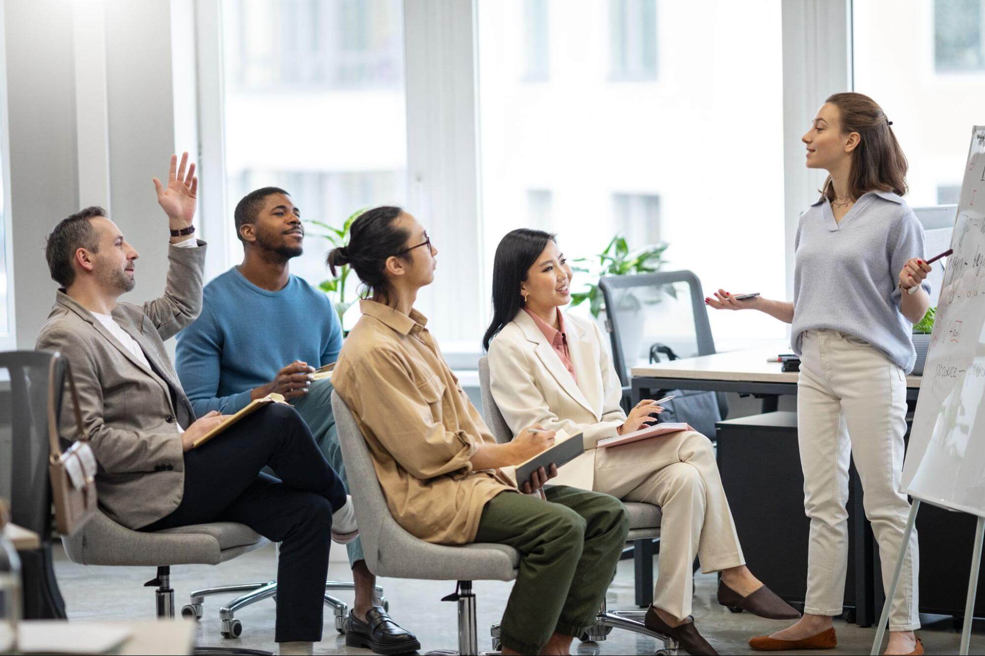 A young woman presents to a diverse group of four in an office, with a whiteboard and an engaged audience. 