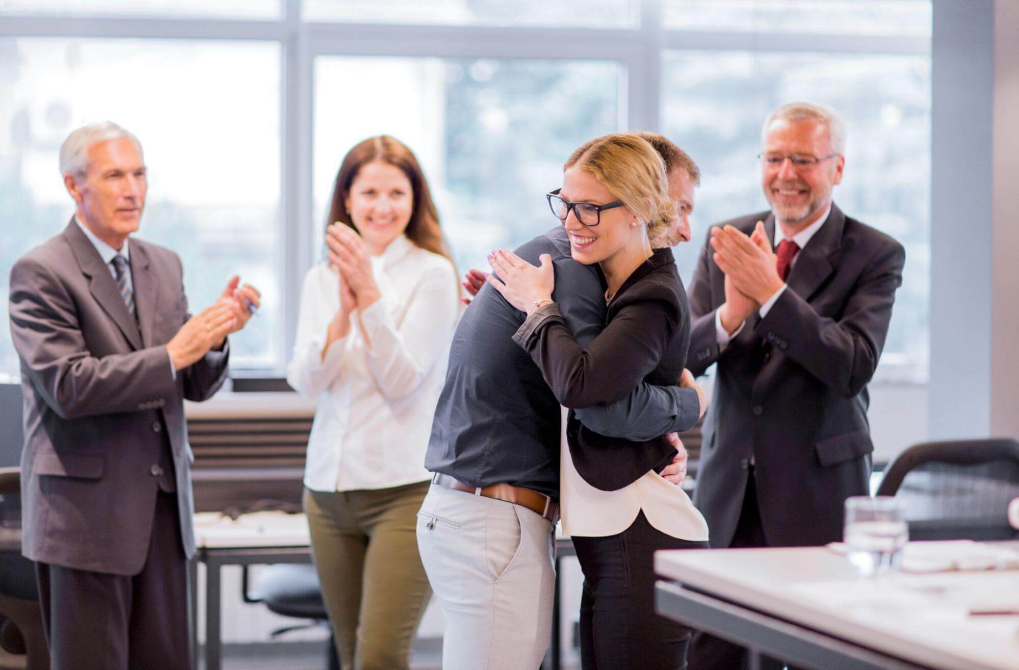 Five professionals in a modern office celebrate a success, with hugs and applause, city view behind.