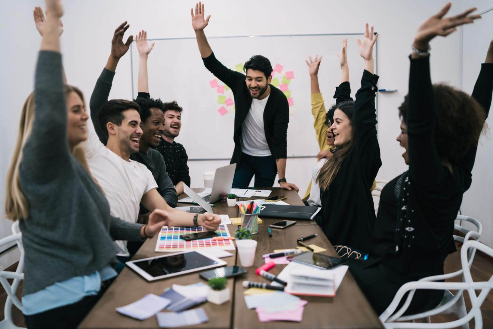 Seven young professionals celebrate a success around a conference table, with laptops, tablets, and papers on the table.