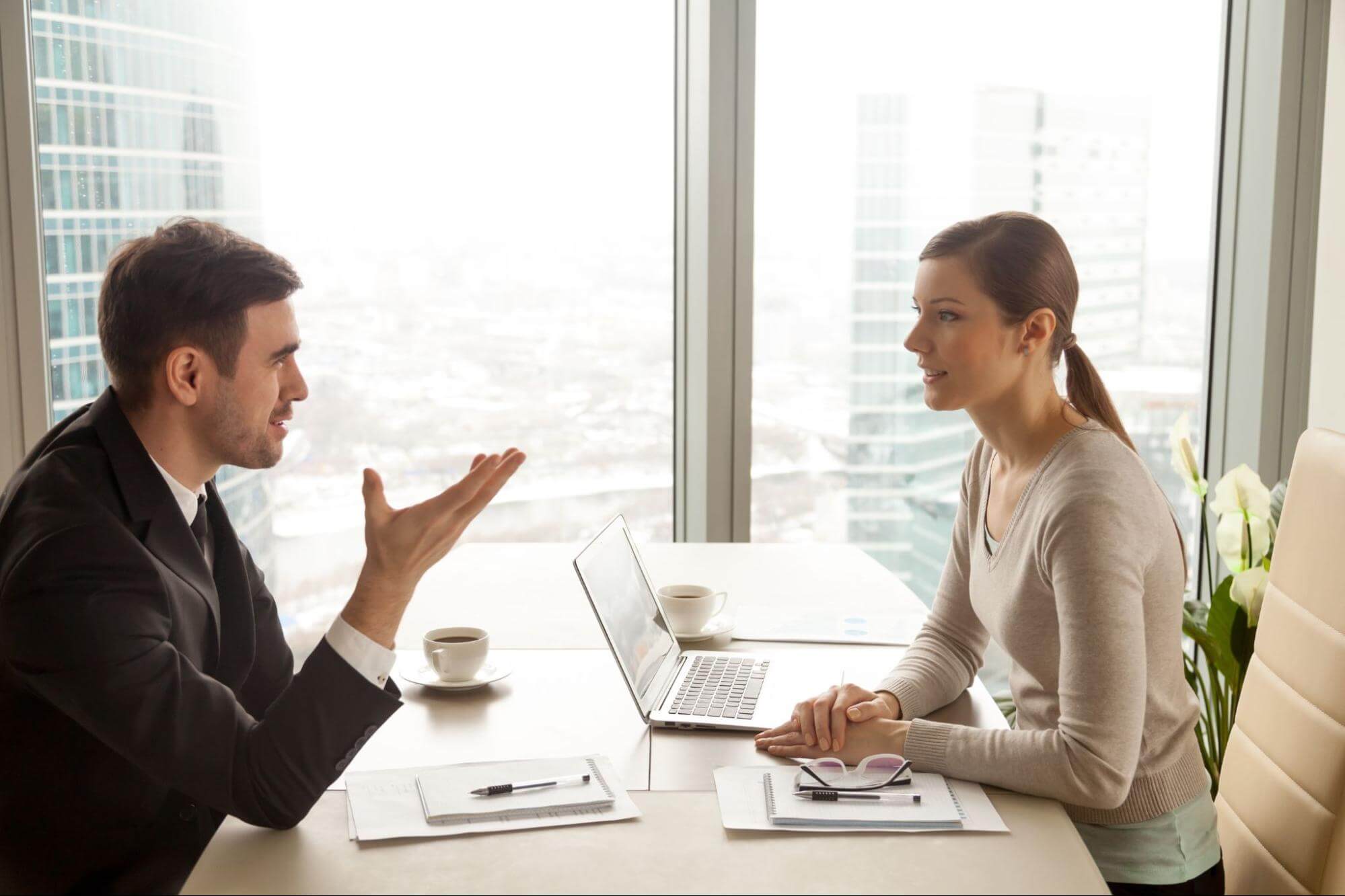 Two young professionals at a desk near a window, discussing with a laptop, coffee, and papers. 