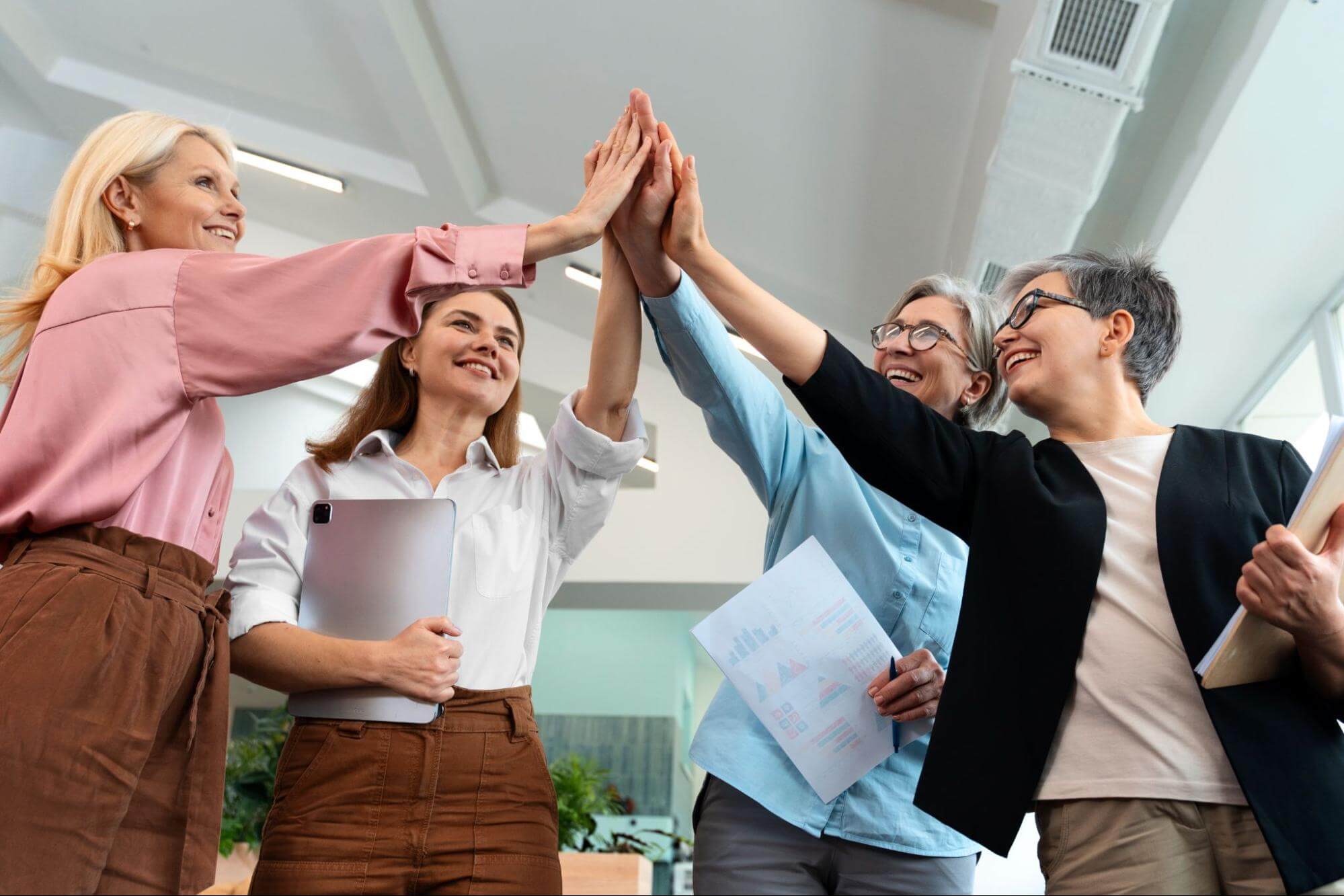 Four professional women of different ages high-five and celebrate success, some holding a tablet and papers. 
