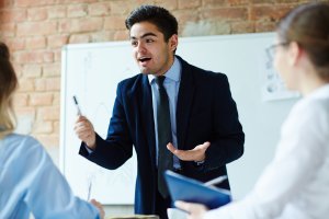 Man in a suit passionately speaks for an executive coaching, gesturing with a pen. Colleagues listen intently. A whiteboard and brick wall are in the background.