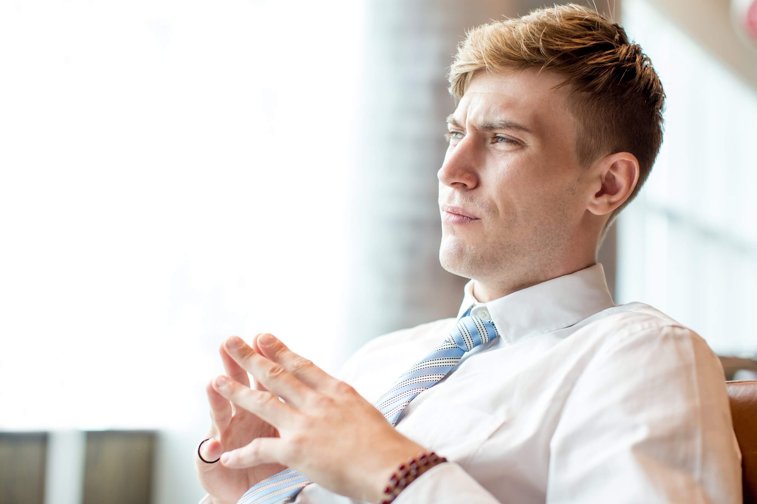 Young man in a white shirt and tie, seated, looking thoughtfully into the distance with clasped hands. Bright, softly lit background.
