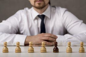 A man in a white shirt and tie sits at a table with six chess pawns, five light and one dark. The image suggests themes of leadership and self-regulation.