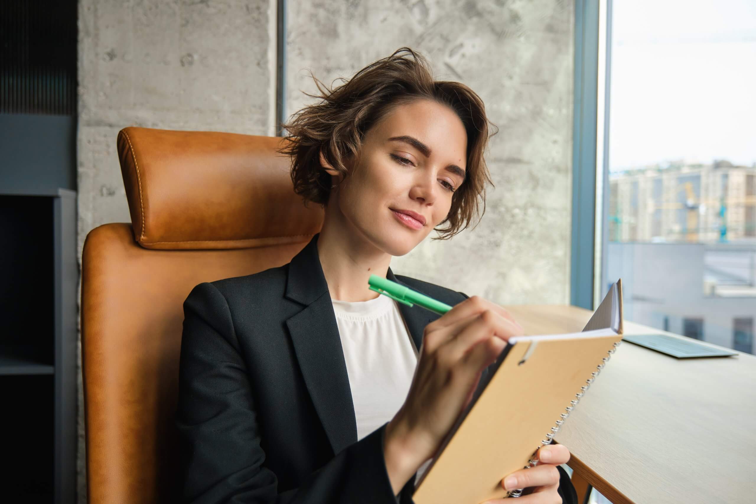 A confident woman in a blazer sits in a leather chair by a window, journaling in a notebook. She looks focused and content.