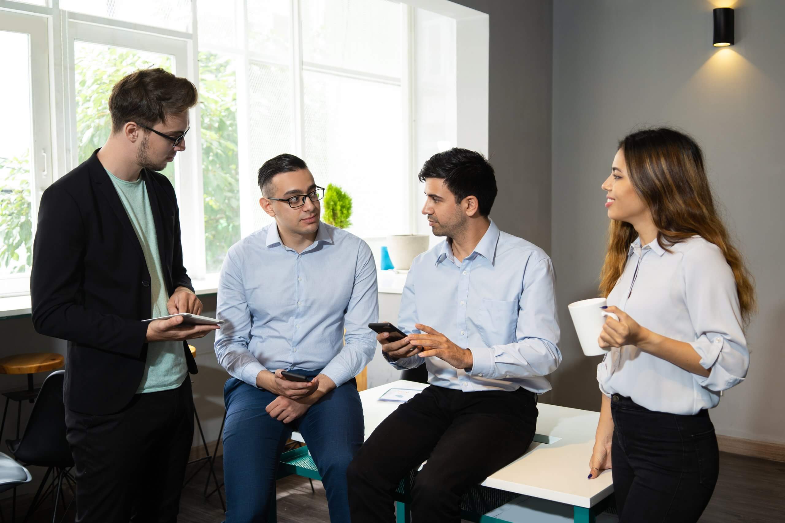Four colleagues in a bright office discuss work. Three men, one holding a tablet, sit on a table while a woman stands with a coffee cup, smiling.
